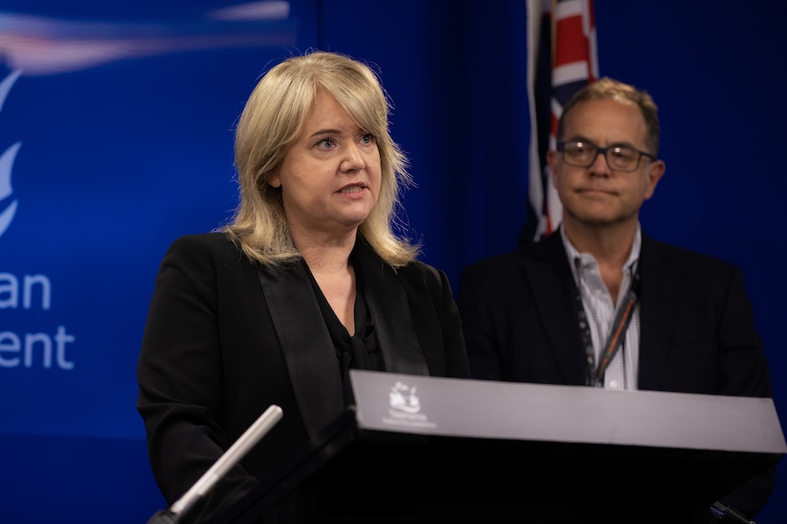 A woman with blonde hair stands at a lectern with a man looking on