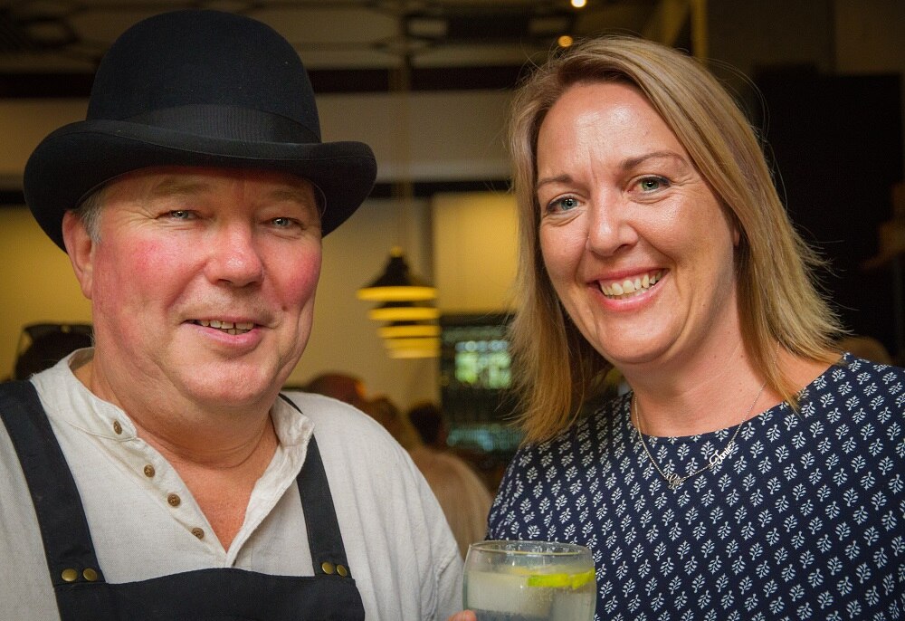 Hombre con sombrero y delantal con mujer de cabello rubio, ambos sonriendo.