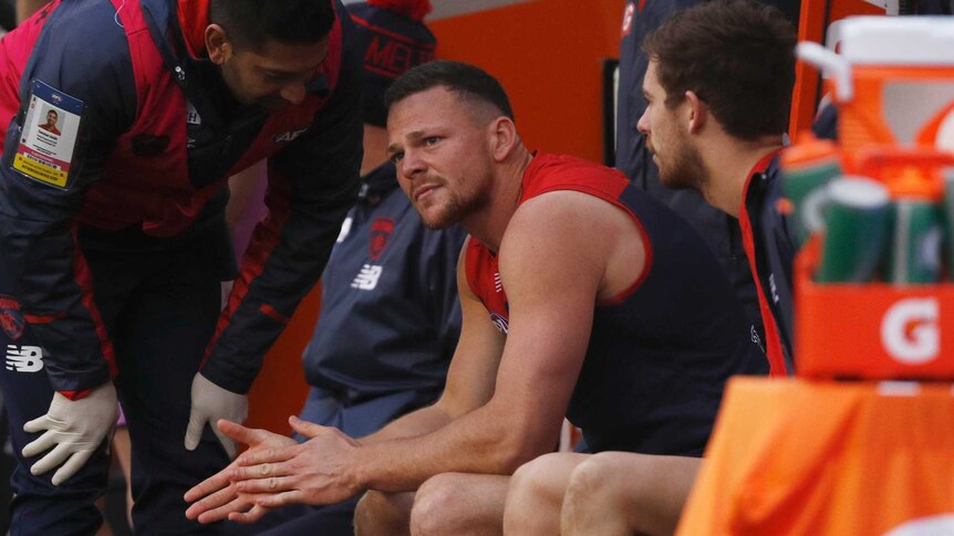 Steven May sits teary-eyed on the Melbourne Demons bench after suffering an injury. A trainer sees to him during the AFL match.
