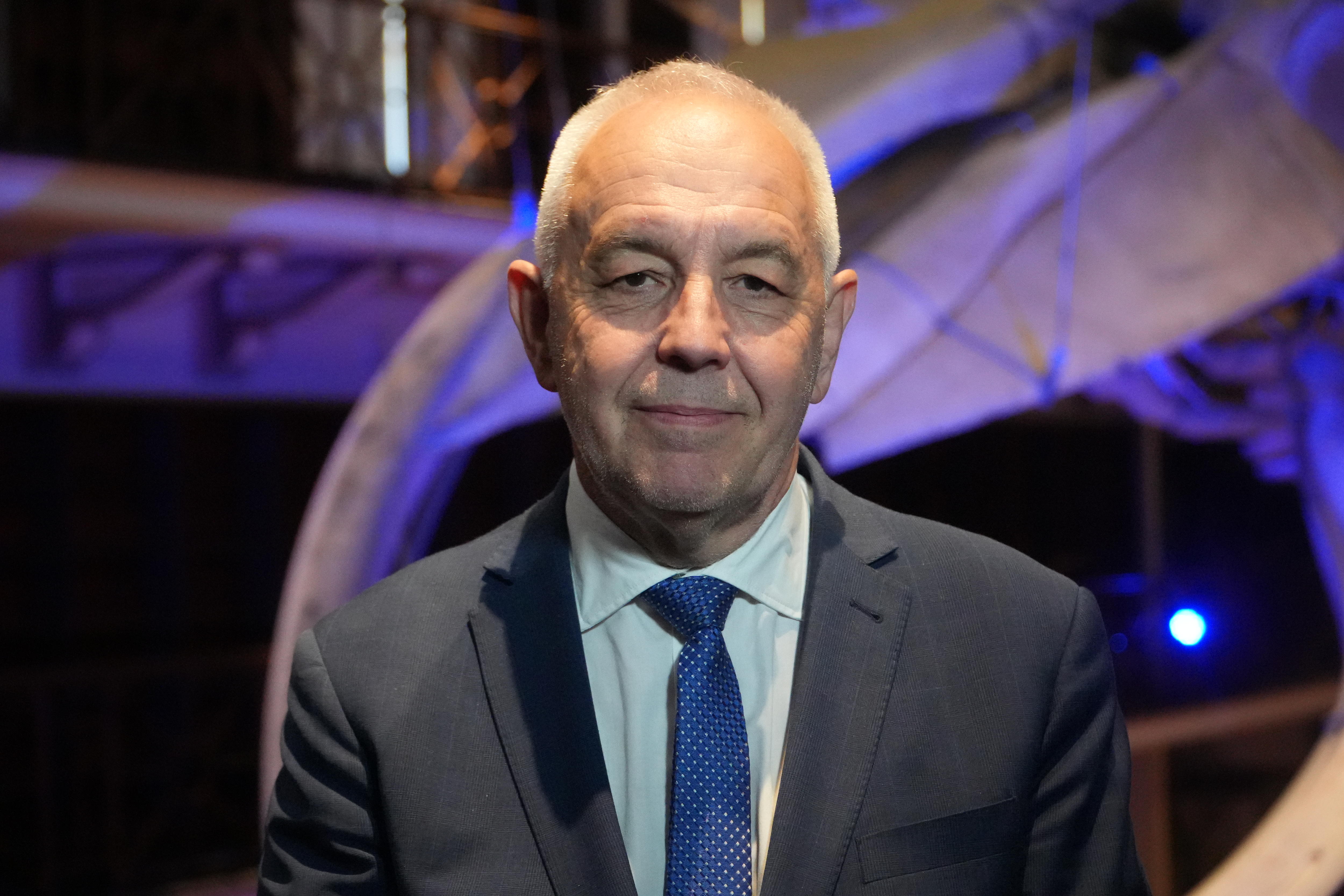 A balding man in a suit and tie named Alec Coles smiles for the camera.