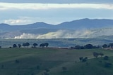 Dust rising off a mine site with green hills in the foreground and background.