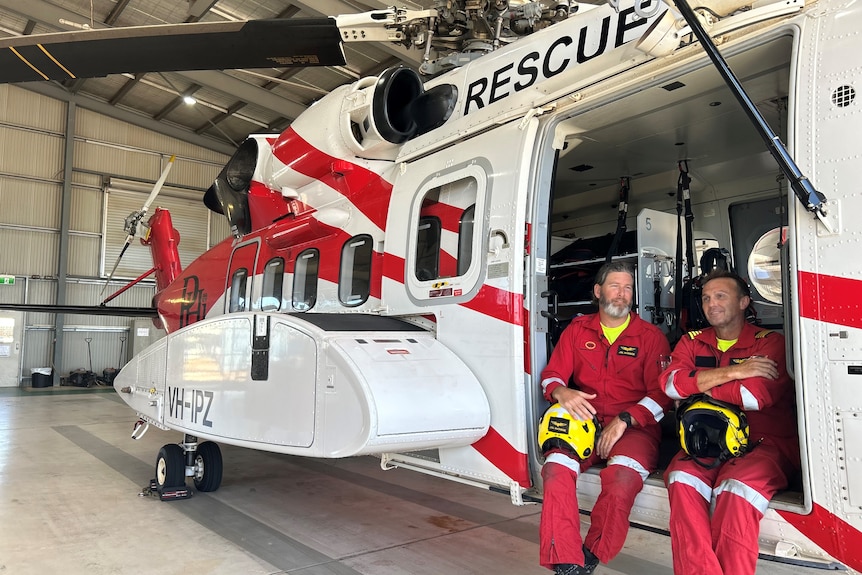 Two men wearing red flight suits sit on a rescue helicopter. 