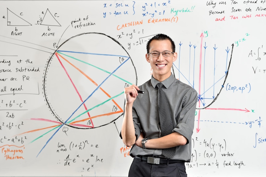 Man wearing dark-coloured shirt, pants and tie stands smiling in front of whiteboard covered in colourful maths formulas.