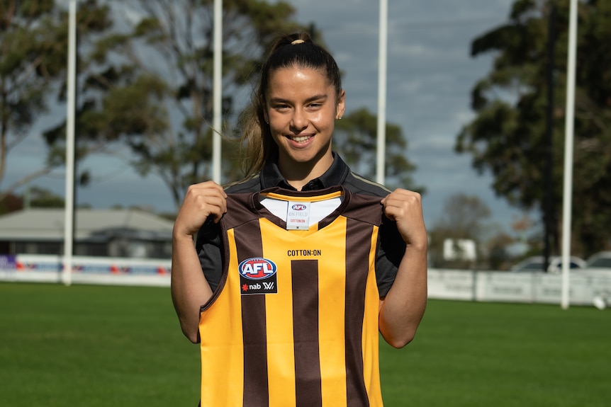 Dominque Carbone hold up a Hawthorn jersey on a football oval.