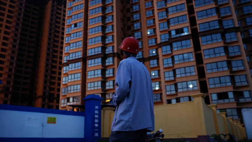 A man in hard hat and blue shirt looks upwards towards an apartment building