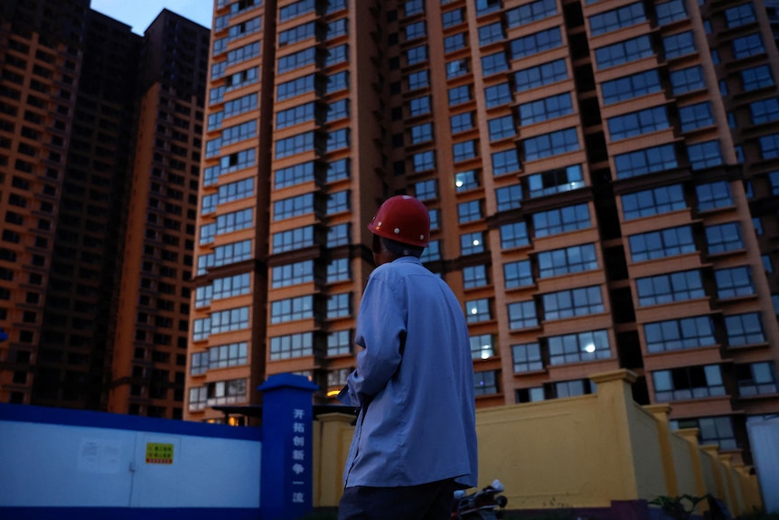 Un hombre con casco y camisa azul mira hacia un edificio de apartamentos