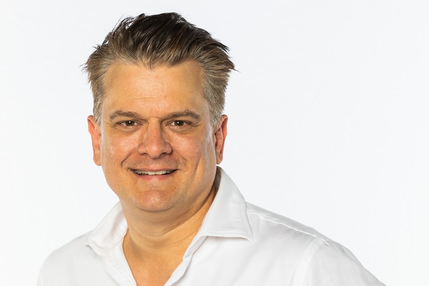 A headshot of a smiling man with short brown hair, wearing a white shirt