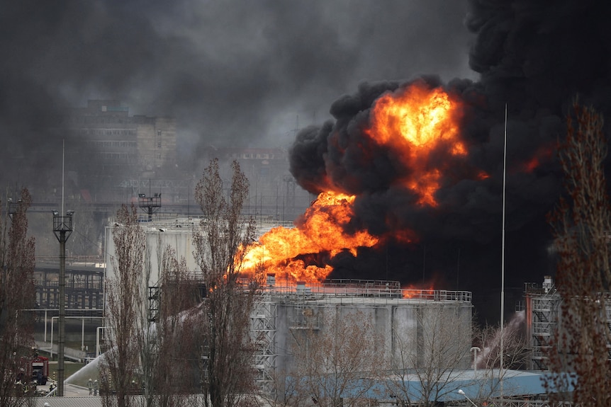 A water jet points upwards towards a fire and black smoke on a large industrial tank.