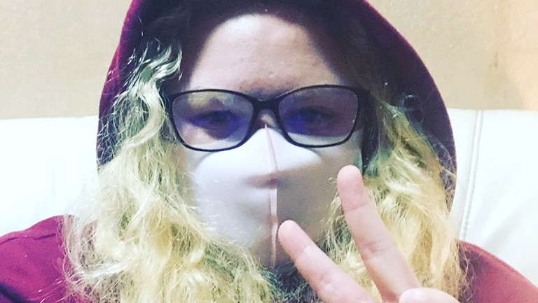 A woman in her 20s with long blonde hair, hat and glasses wears a mask across her nose and mouth.