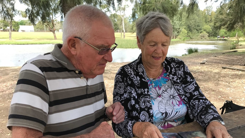 An elderly couple looking through a photo album in a park with a lake and trees in the background.