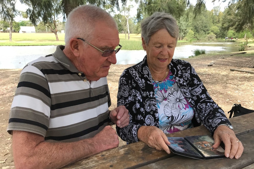 An elderly couple looking through a photo album in a park with a lake and trees in the background.