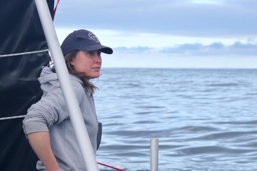 A woman wearing a baseball cap stands on a boat and looks out the the ocean.