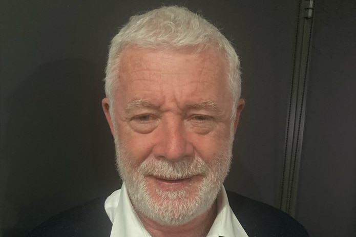 A man with white hair and a beard wearing a white colared shirt and black jumper