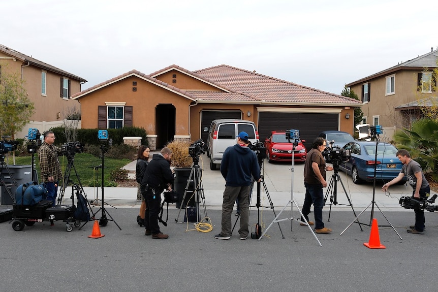 Reporters stand outside a single-story brick house.