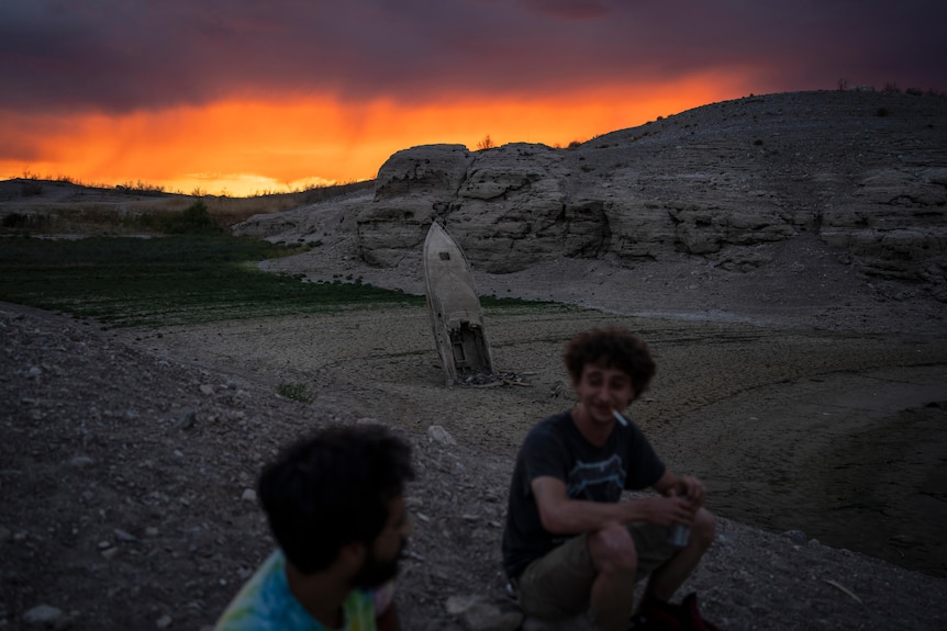 Men smoke at dusk on lake bed where boat behind them sits upright. On horizon sunset blazes yellow, red and purple. 