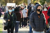 People wearing masks wait in line on a sunny day.