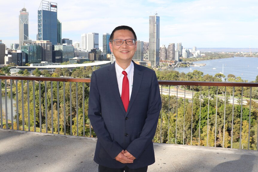A man with glasses, a suit and a red tie smiles at the camera.