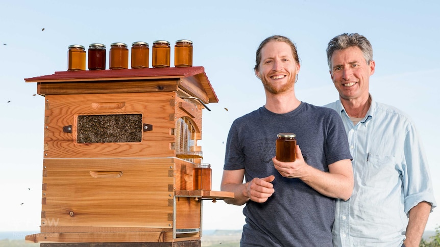 Stuart Anderson and his son Cedar stand next to a beehive, holding honey and smiling.