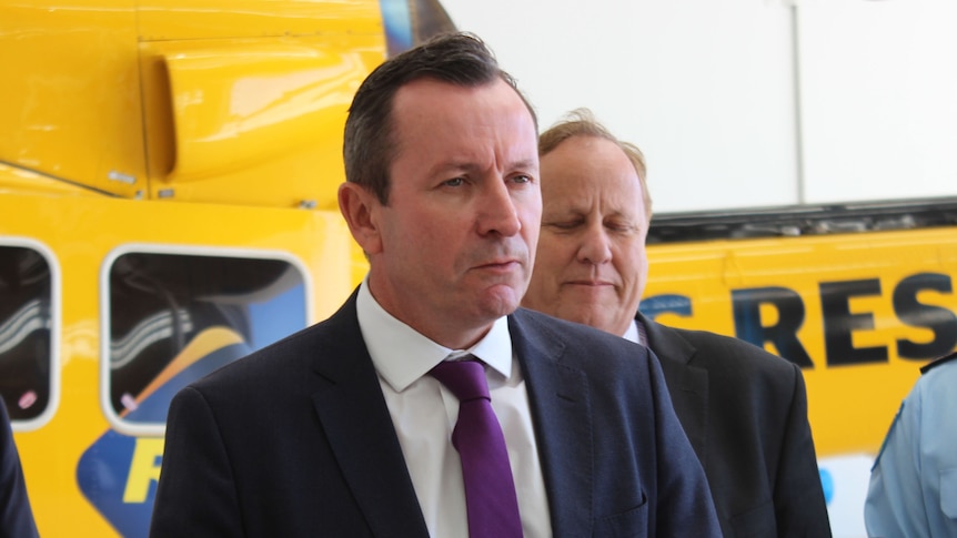 The WA Premier Mark McGowan stands in front of a yellow rescue helicopter while being interviewed by journalists.