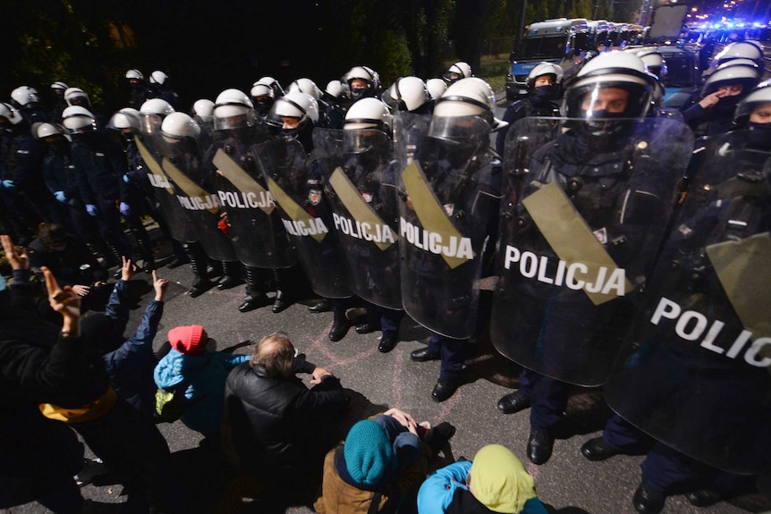 A police cordon with riot shields stands in a line in front of seated protesters, near the house of a senior politician.