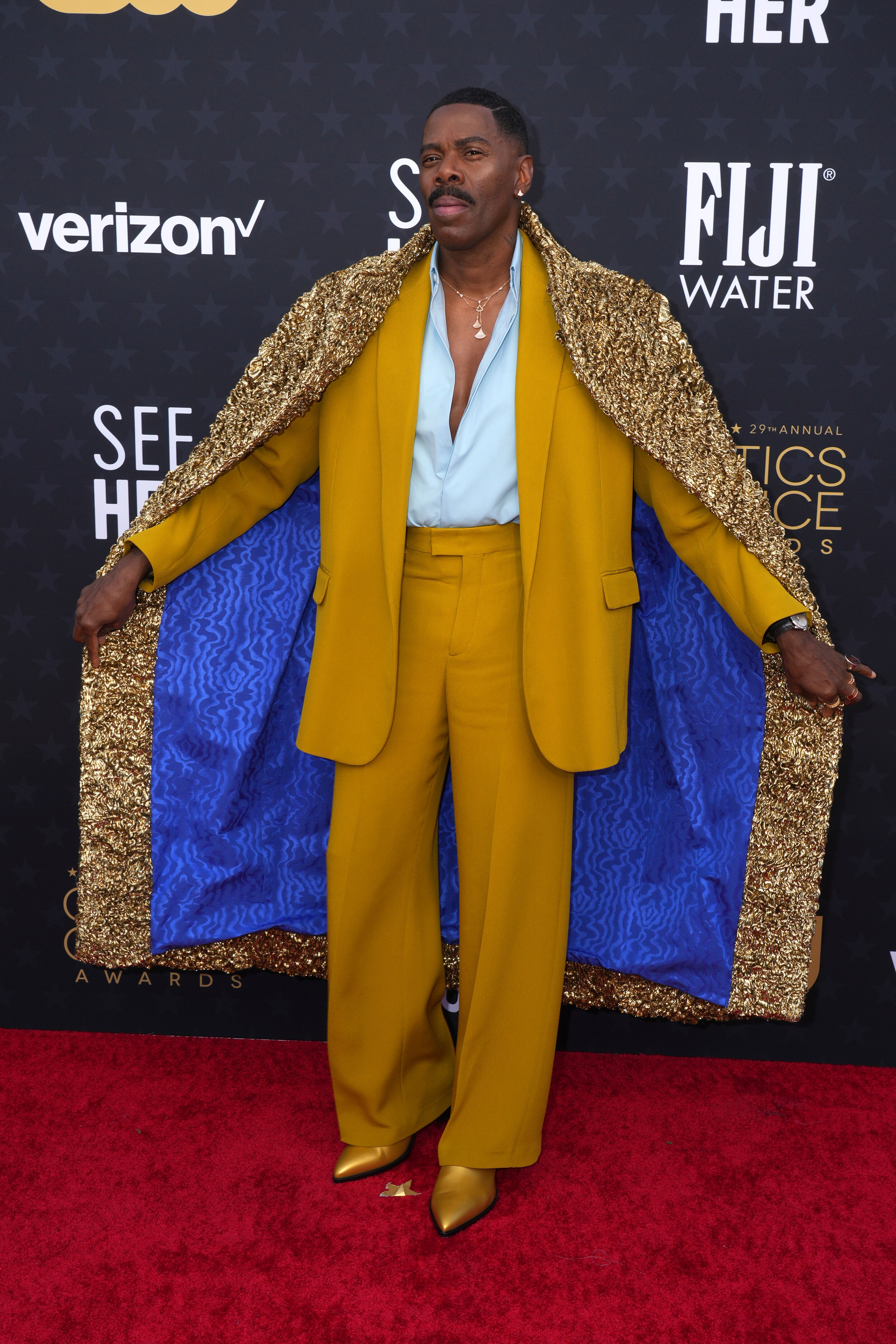 Colman Domingo vistiendo un traje dorado con una camisa blanca y fluida y una capa con detalles metálicos dorados en el exterior y forro azul.