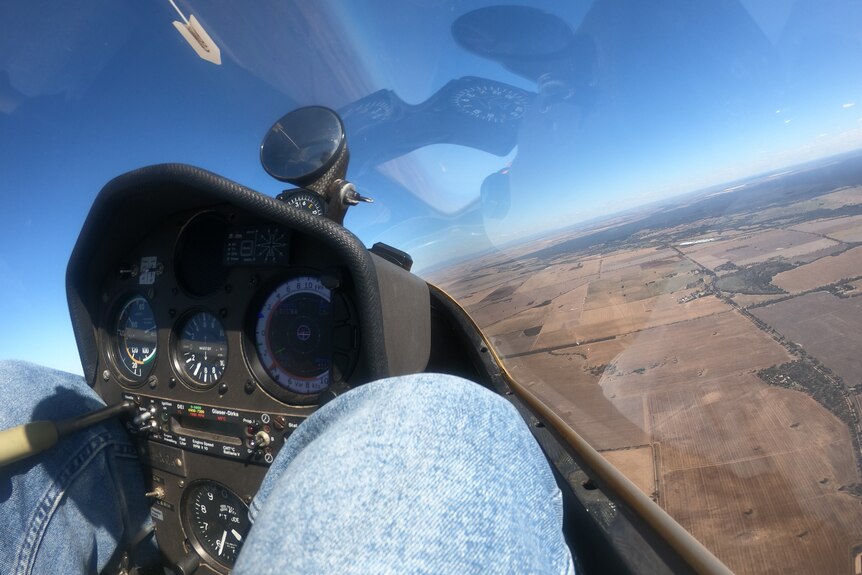 A close up of a pair of jeans-clad legs clutching a glider's flight instrument inside a cockpit. Blue sky and paddocks seen.