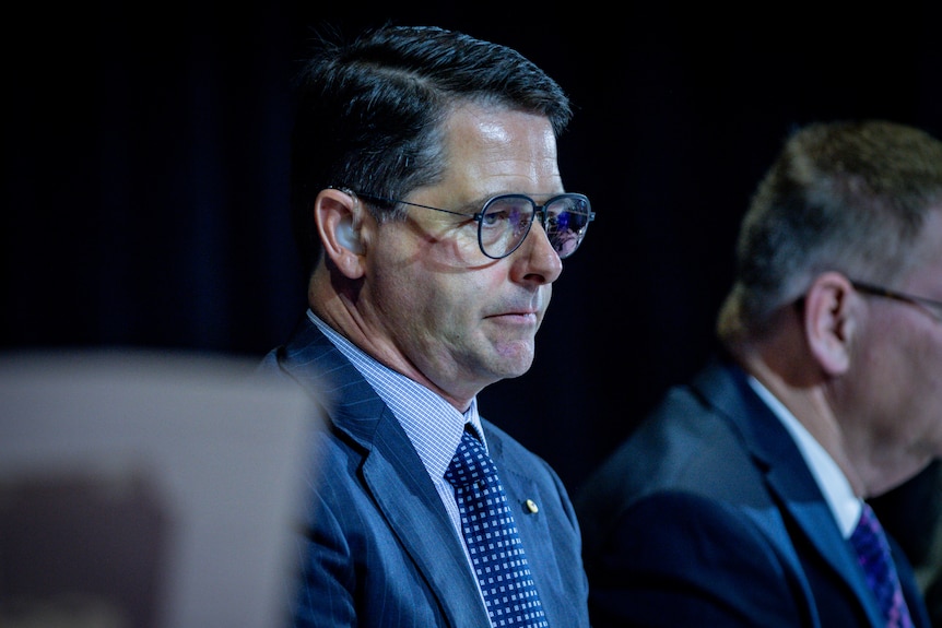 A white man with short black hair and a navy suit. He is wearing a hearing aid and sitting on a stage