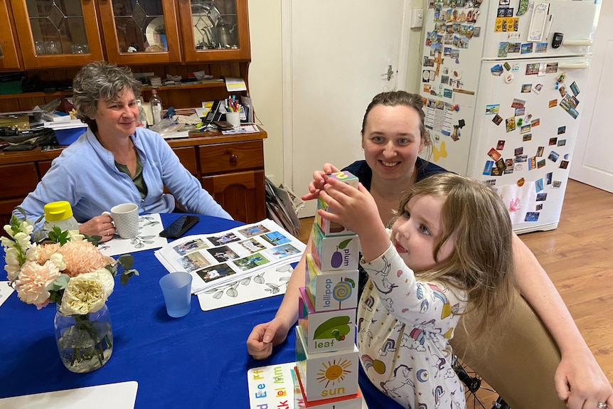 A child builds a tower of blocks as her mum and grandma watch, sitting at a kitchen table