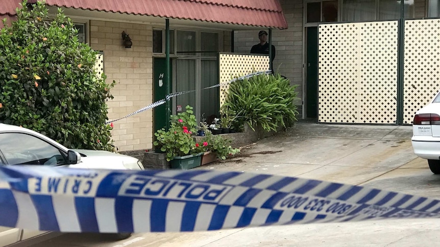 A driveway is cordoned off with blue and white police tape as an officer stands guard.