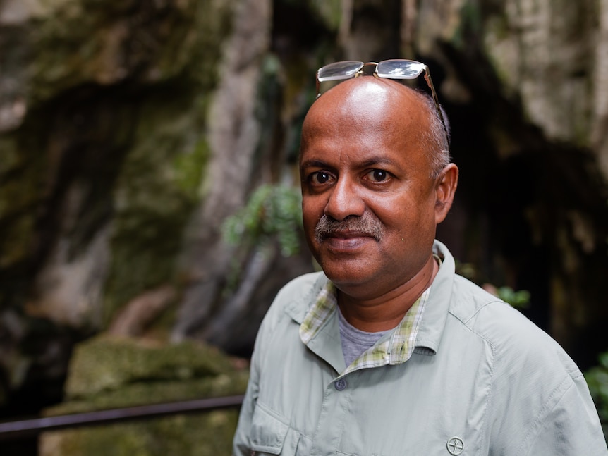 A man looks at the camera, with glasses resting on his head, caves and moss in background