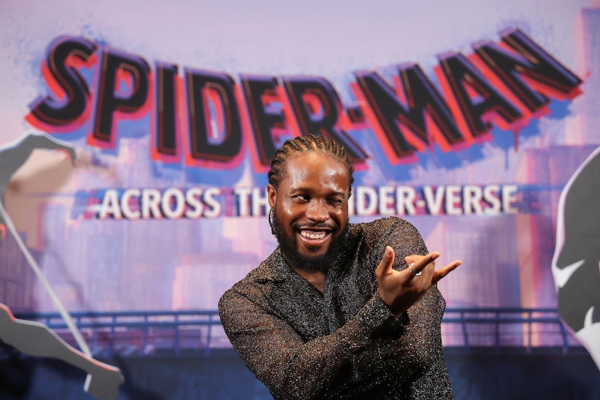 A man smiling, with his hand posed like spider-man in front of a promotional backdrop. 