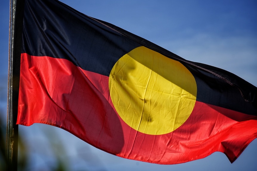 An Indigenous flag flaps in the wind against a blue sky background