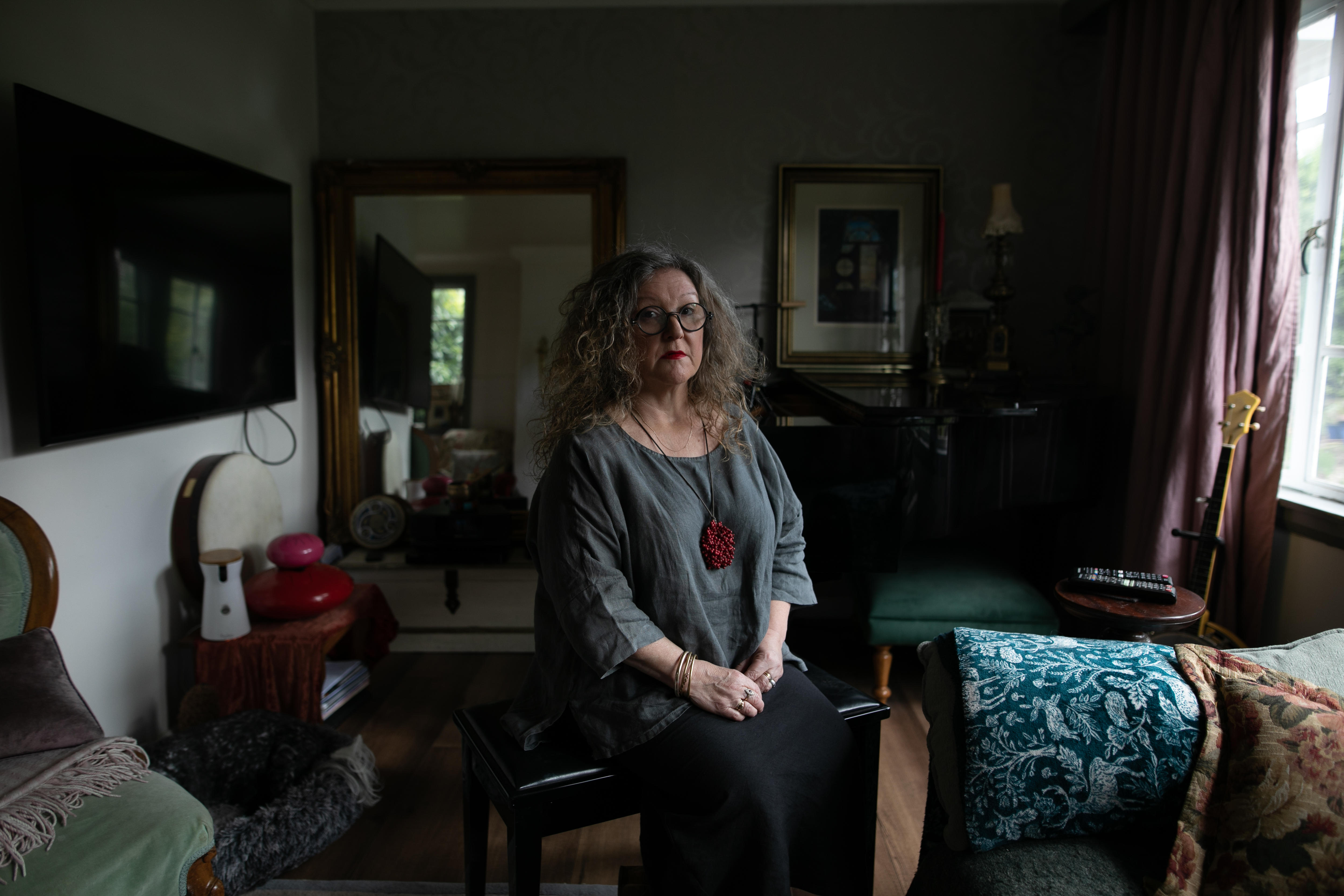 A woman wearing loose grey linen top sits on a stool in the middle of a cluttered living room, looking solemn at camera