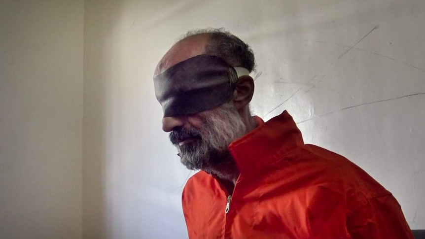 Older man with grey beard sits in orange prison uniform wearing brown blindfold in dim lit room