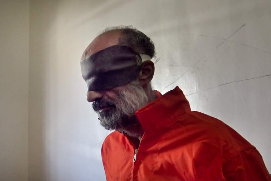 Older man with grey beard sits in orange prison uniform wearing brown blindfold in dim lit room