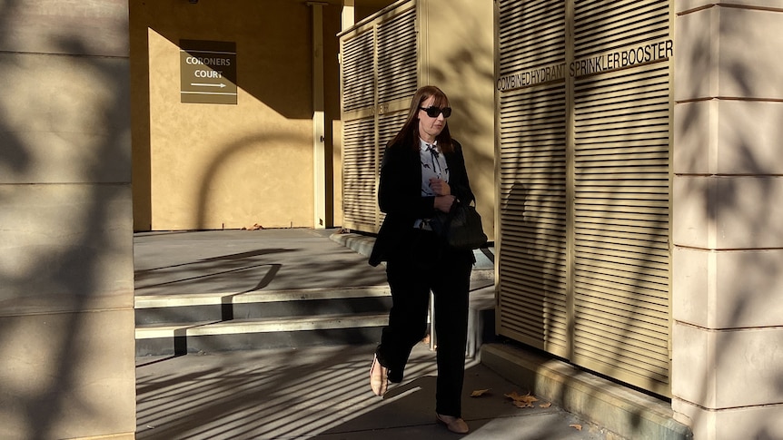 A woman in black pants and blazer with brown hair walks out of coroners court.