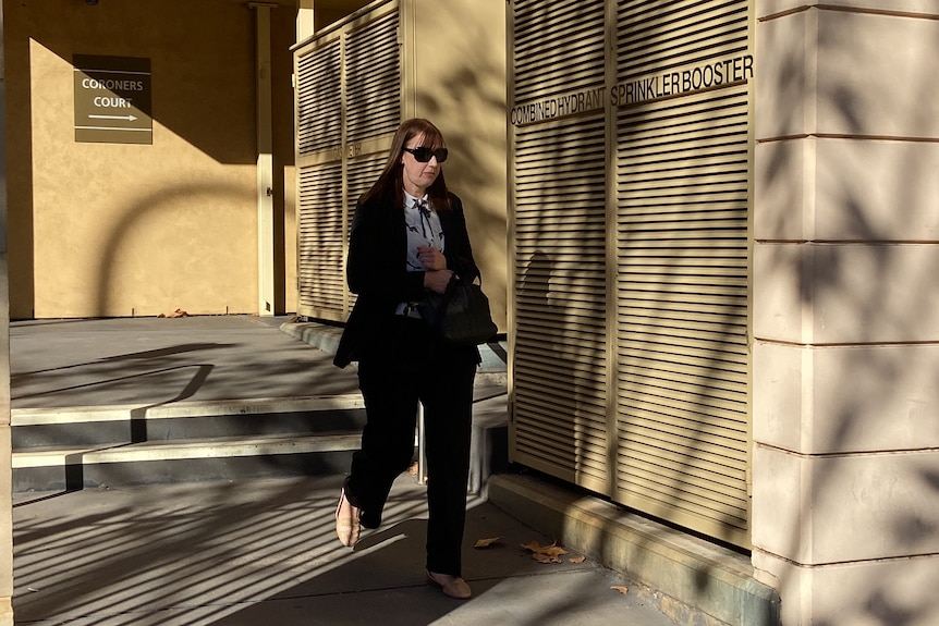 A woman in black pants and blazer with brown hair walks out of coroners court.