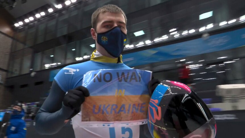 A Winter Olympian in blue and white and black face mask holds up 'No Way in Ukraine' sign at indoor venue