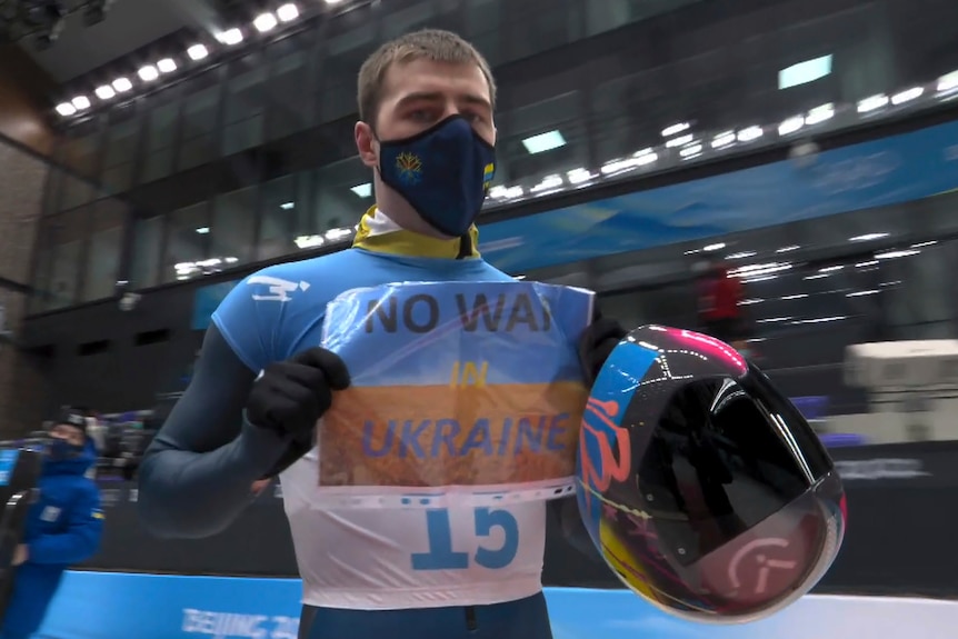 A Winter Olympian in a blue and white and black mask holds up the sign 