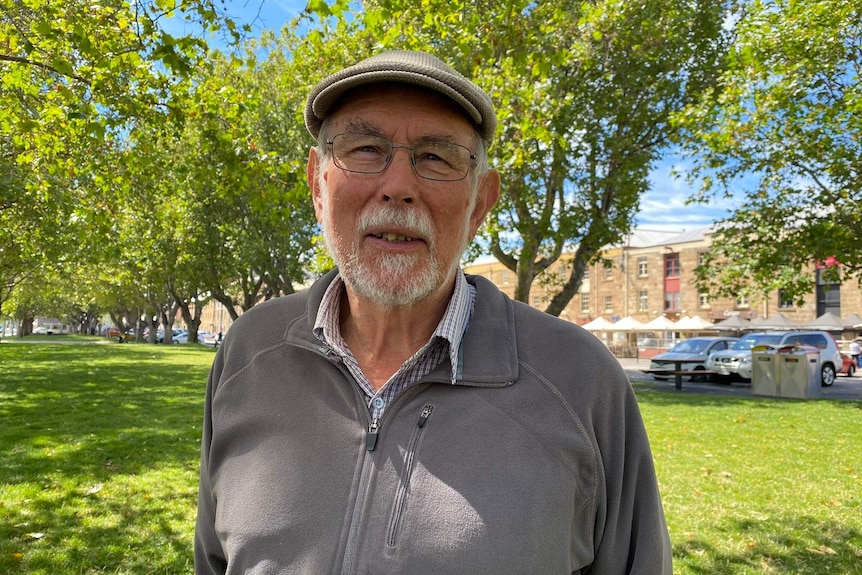 A older man wearing a baker boy hat and glasses stands on a lawn in front of trees.