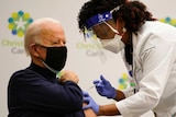 Joe Biden sits on a chair, wearing a black mask, as a nurse leans over, pressing the needle into his arm.