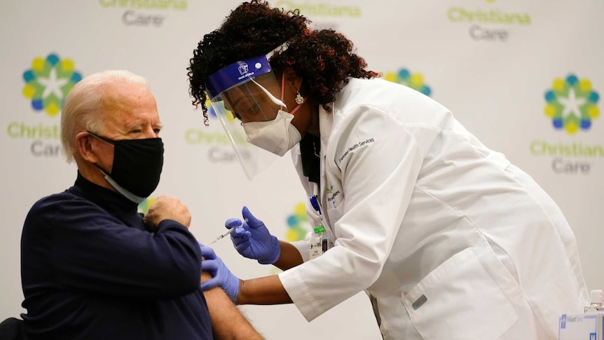 Joe Biden sits on a chair, wearing a black mask, as a nurse leans over, pressing the needle into his arm.