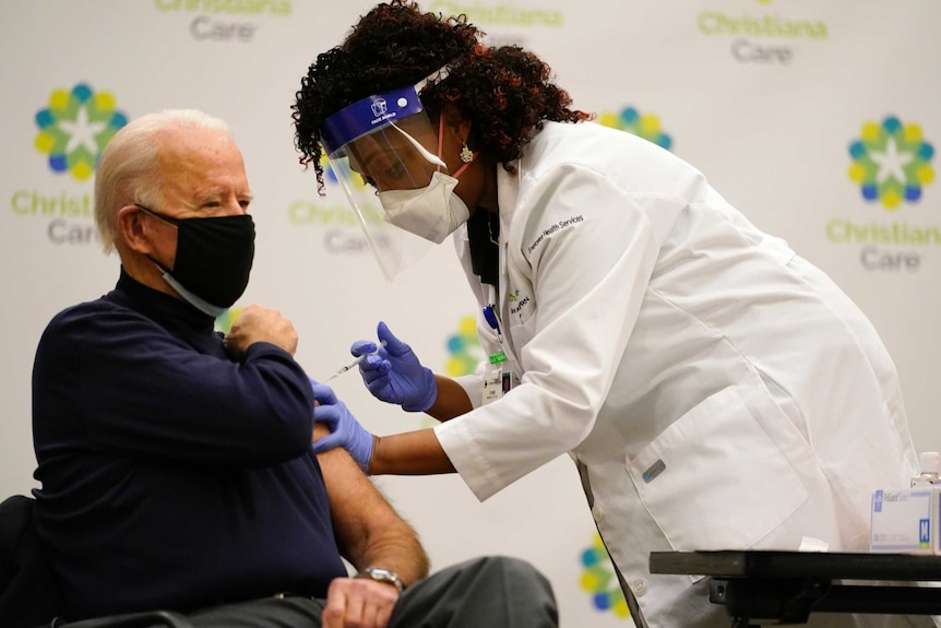 Joe Biden sits on a chair, wearing a black mask, as a nurse leans over, pressing the needle into his arm.