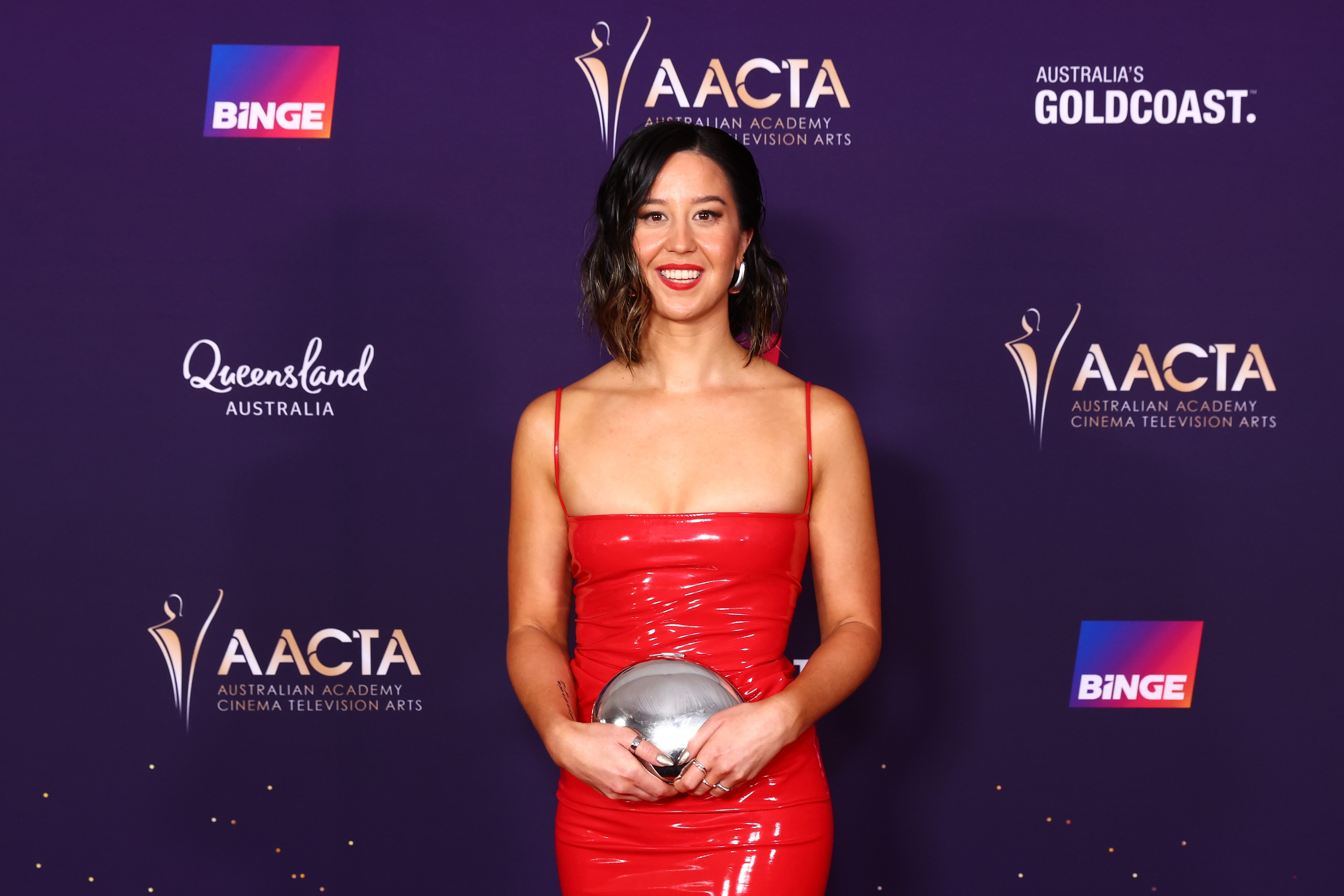 Nina Oyama wears a red, spaghetti strap latex dress with a silver metal purse, silver hoop earrings and a red lip