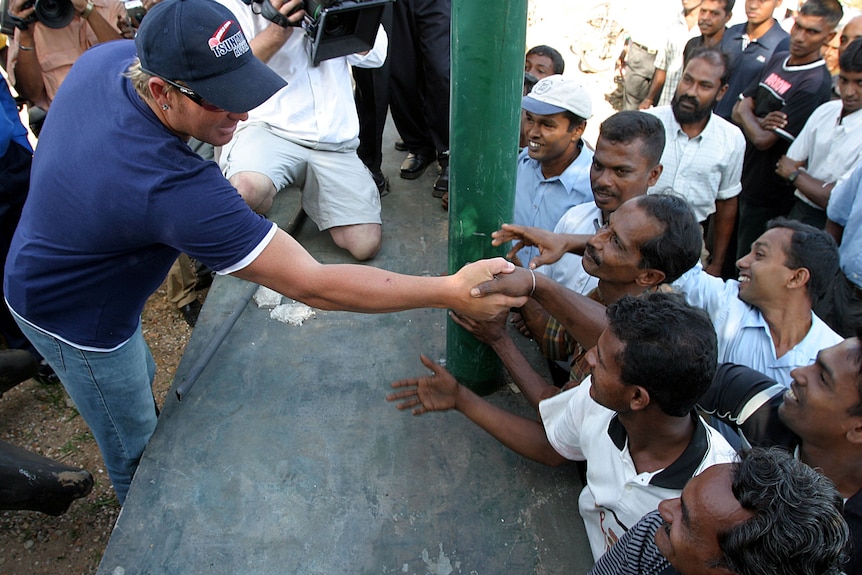 Shane Warne, wearing a blue shirt and cap, reaches out to shake hands with Sri Lankan fans across a barrier