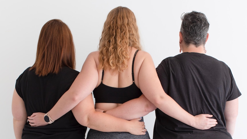 Three women, visible from behind, are linking arms as they show people their bodies. 