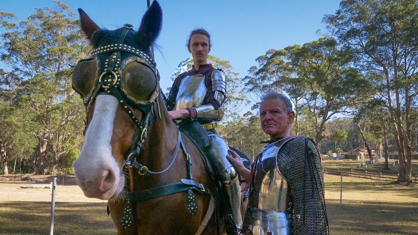 Two men dressed as knights - one mounted and the other standing beside - in armour with trees in the background.