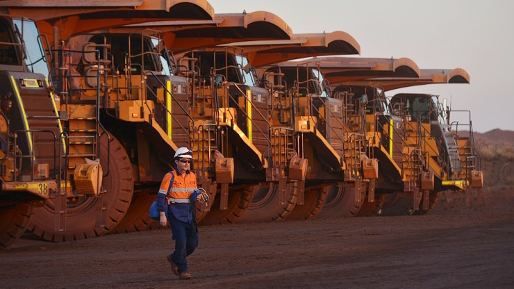 A worker wearing a hardhat and hi-wis clothing walks in front of a row of trucks at ConsMin's Woodie Woodie mine in the Pilbara.