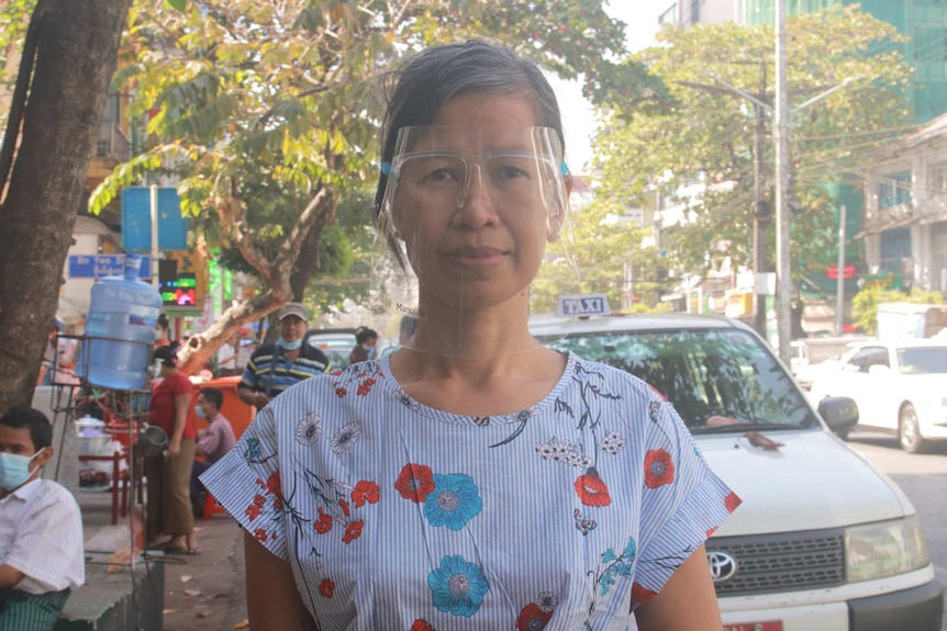 A woman with black hair wears glasses and a face shield as she stands on an empty street.