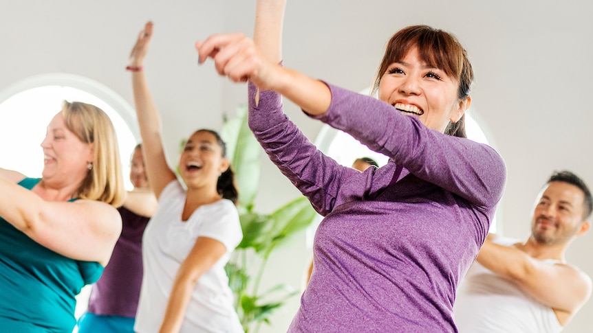 A smiling woman in a purple top holds her hands up high and dances with a group of people